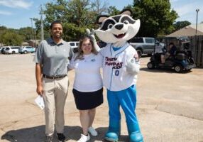 A man and a woman dressed for a golf tournament, standing with a large raccoon mascot of the Rocket City Trash Pandas. They are standing in a parking lot and smiling.